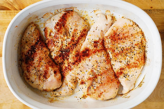 4 chicken breasts in a white casserole dish are seasoned with spices