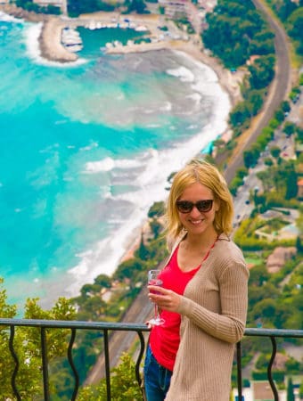 a woman dressed in red top and jeans - standing with a wine glass overlooking the spectacular sea view from the top of village Eze.