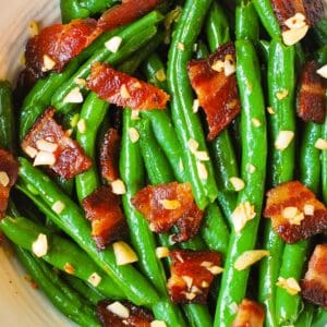 Bright green beans tossed with crispy bacon pieces and minced garlic, all glistening with oil, fill a white bowl.