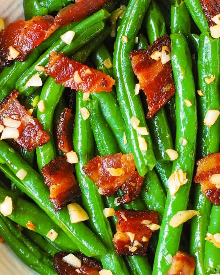 Bright green beans tossed with crispy bacon pieces and minced garlic, all glistening with oil, fill a white bowl.