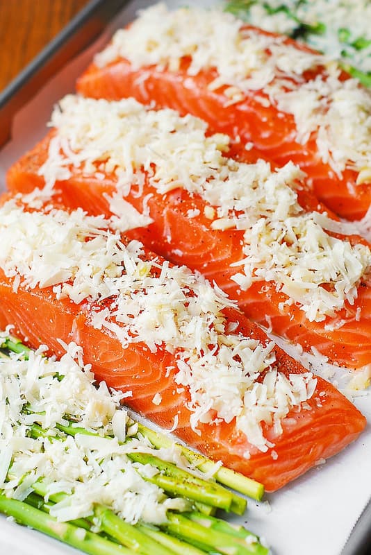 raw salmon sprinkled with shredded Parmesan cheese and raw asparagus topped with shredded Parmesan cheese on a parchment paper on baking sheet