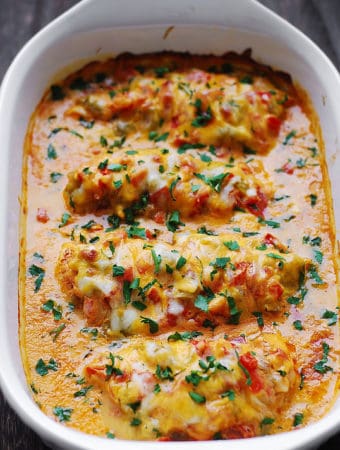 Queso Baked Chicken - chicken spiced up with Southwestern spices and smothered with chopped tomatoes, green chiles, Queso sauce, topped with Cheddar - in a white casserole dish.