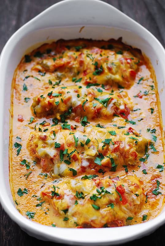 Queso Baked Chicken - chicken spiced up with Southwestern spices and smothered with chopped tomatoes, green chiles, Queso sauce, topped with Cheddar - in a white casserole dish.