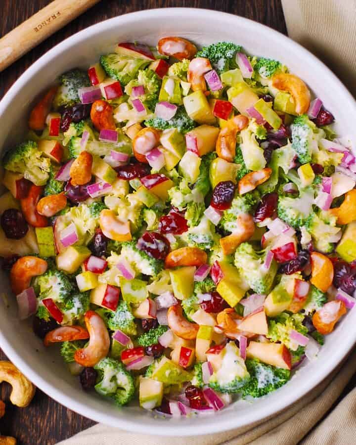 A bowl of colorful salad with broccoli, diced red onions, red bell peppers, apple chunks, dried cranberries, and cashew nuts, all mixed with a creamy dressing. Cashews are scattered nearby on a wooden table.