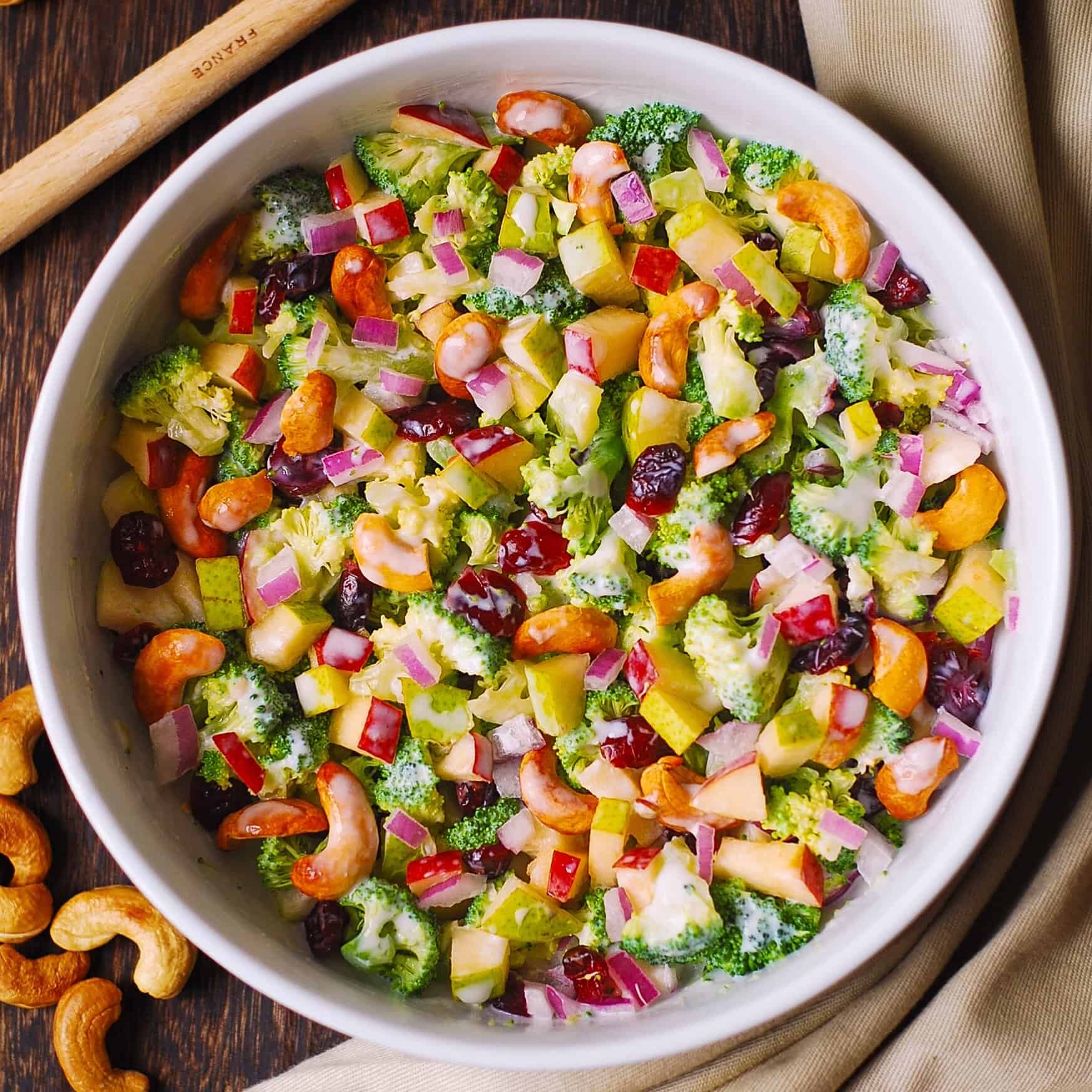 A bowl of colorful salad with broccoli, diced red onions, red bell peppers, apple chunks, dried cranberries, and cashew nuts, all mixed with a creamy dressing. Cashews are scattered nearby on a wooden table.