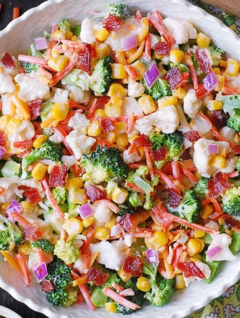 Creamy Broccoli, Cauliflower, Corn, Bacon Salad with Sliced Carrots, Diced Red Onions, and shredded Sharp Cheddar Cheese in a white bowl