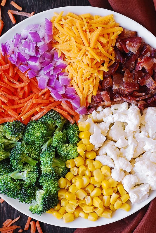 sections of salad ingredients on a white plate, clock-wise: shredded cheddar cheese, chopped cooked bacon, chopped broccoli florets, corn kernels, chopped broccoli, sliced carrots, chopped red onions