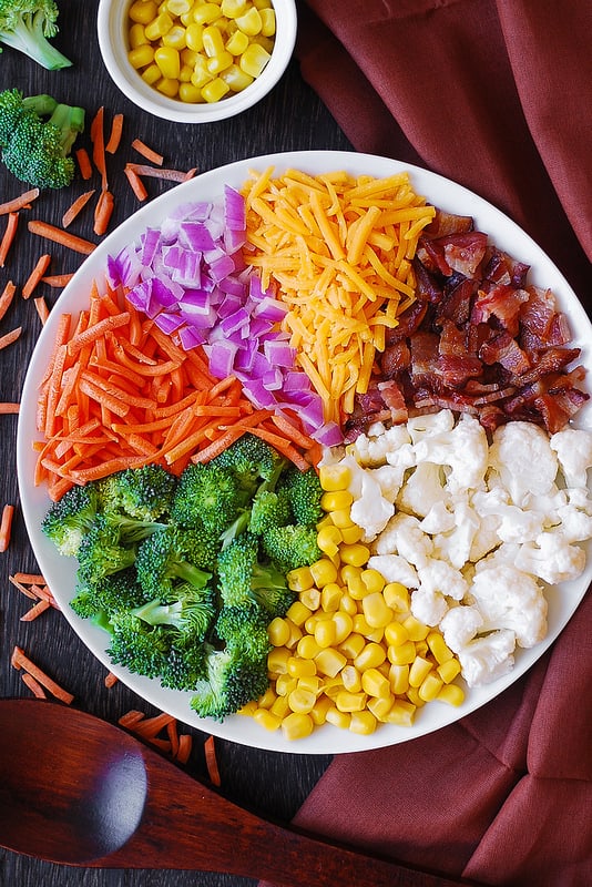 sections of salad ingredients on a white plate, clock-wise: shredded cheddar cheese, chopped cooked bacon, chopped broccoli florets, corn kernels, chopped broccoli, sliced carrots, chopped red onions