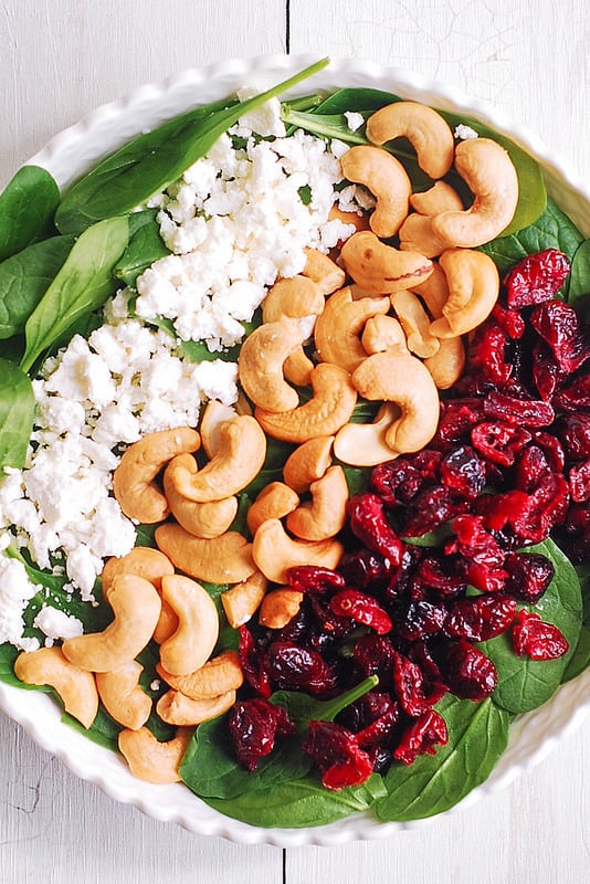 Spinach in the salad bowl with layers of ingredients on top: crumbled goat cheese, cashews, and dried cranberries