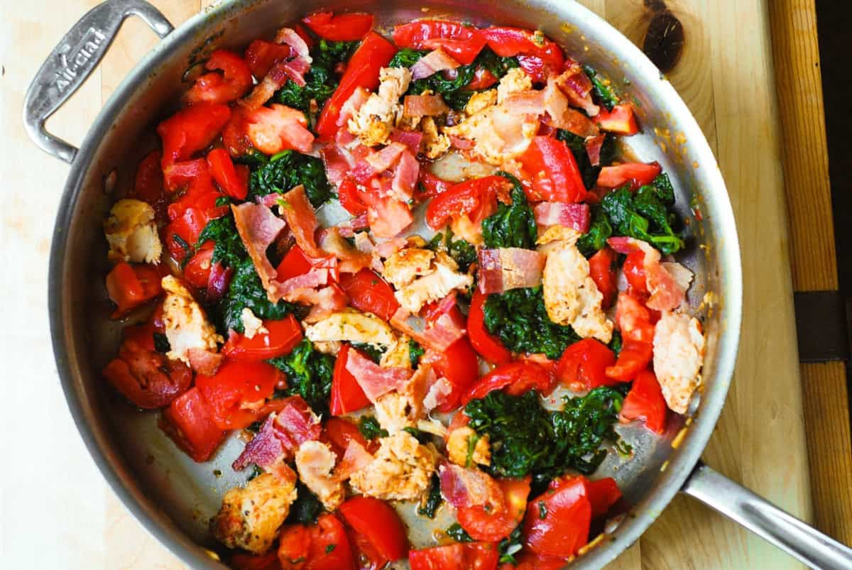 Cooked vegetables (tomatoes, spinach), bacon, and chicken in a stainless steel pan.