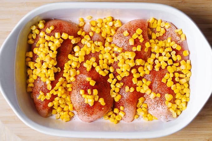 4 raw chicken breasts topped with chili powder and corn kernels in a white casserole dish
