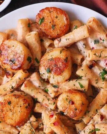 A plate of creamy pasta topped with seared scallops, garnished with chopped herbs and red pepper flakes.