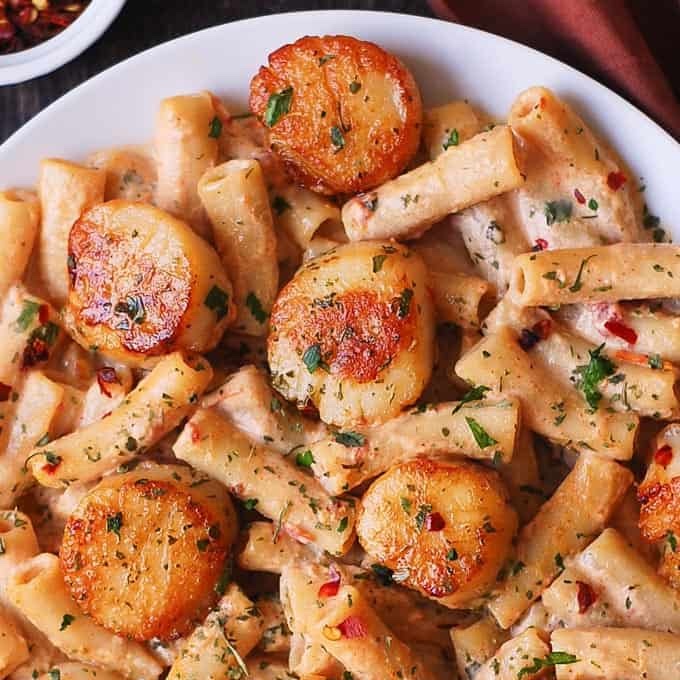 A plate of creamy pasta topped with seared scallops, garnished with chopped herbs and red pepper flakes.
