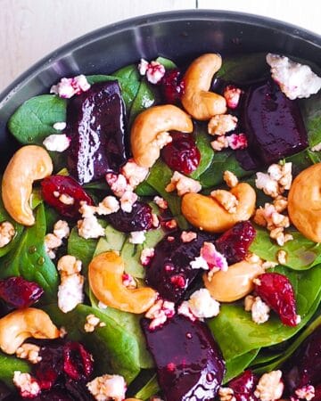 A close-up of a vibrant beet salad featuring fresh spinach leaves, roasted beetroot chunks, cashews, dried cranberries, and crumbled cheese in a black bowl.