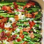 Roasted asparagus spears in a baking dish, topped with crispy bacon pieces and melted cheese or shredded parmesan, with a sprinkle of minced garlic visible.