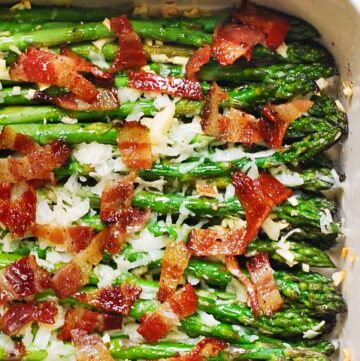 Roasted asparagus spears in a baking dish, topped with crispy bacon pieces and melted cheese or shredded parmesan, with a sprinkle of minced garlic visible.