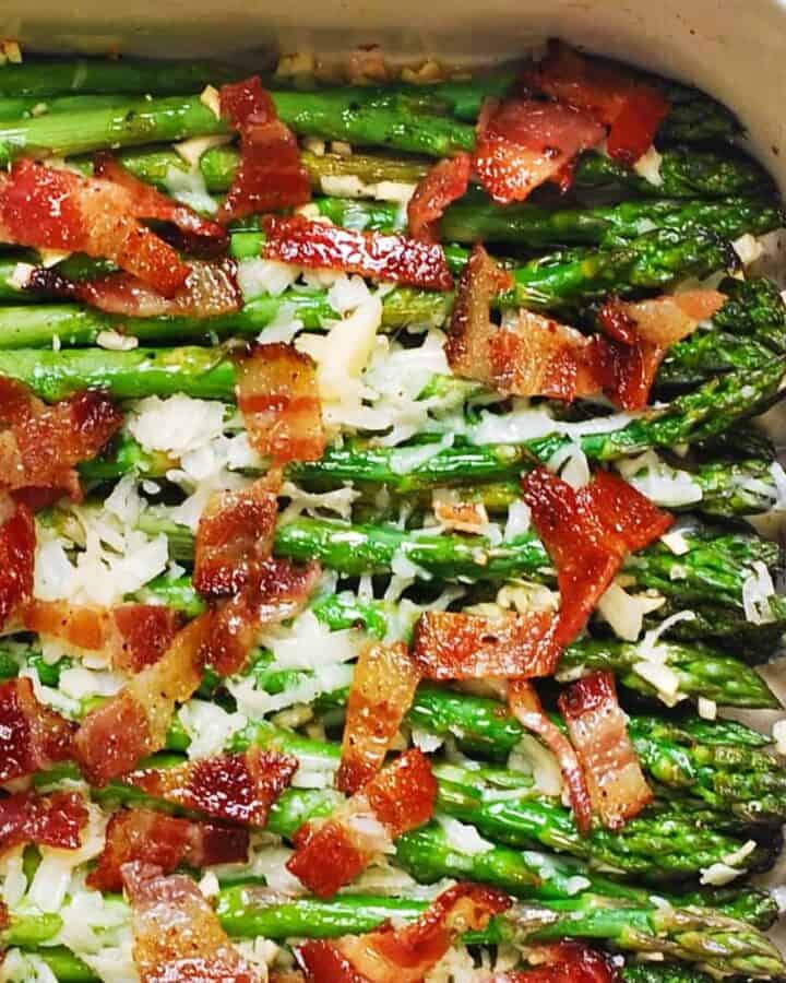 Roasted asparagus spears in a baking dish, topped with crispy bacon pieces and melted cheese or shredded parmesan, with a sprinkle of minced garlic visible.