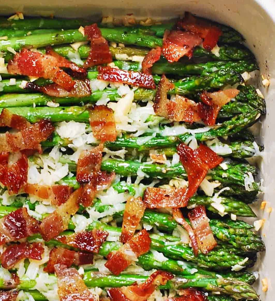 Roasted asparagus spears in a baking dish, topped with crispy bacon pieces and melted cheese or shredded parmesan, with a sprinkle of minced garlic visible.