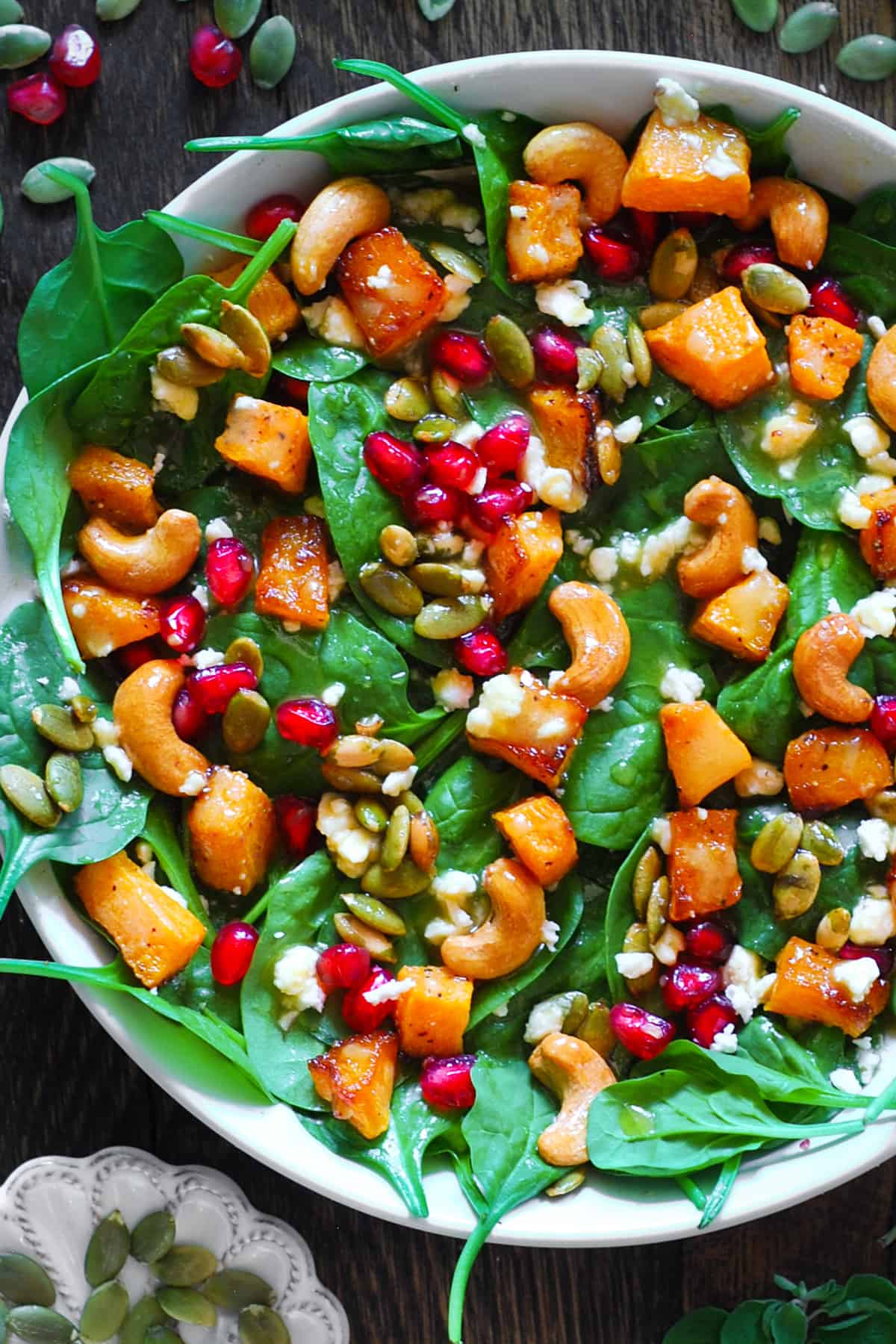 butternut squash salad with baby spinach, roasted cashews, pumpkin seeds, feta cheese, and pomegranate seeds in a white bowl.