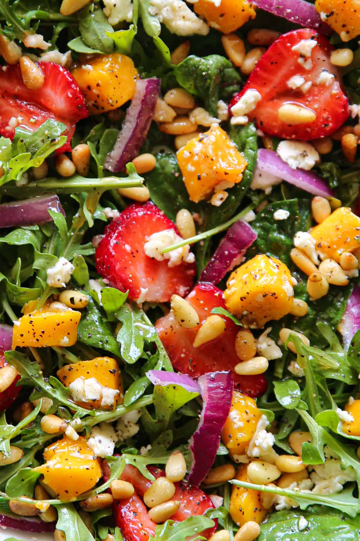 Strawberries and mango salad with fresh greens (arugula and spinach), sliced red onion, feta cheese, and pine nuts in a lemon honey mustard dressing - close-up photo.