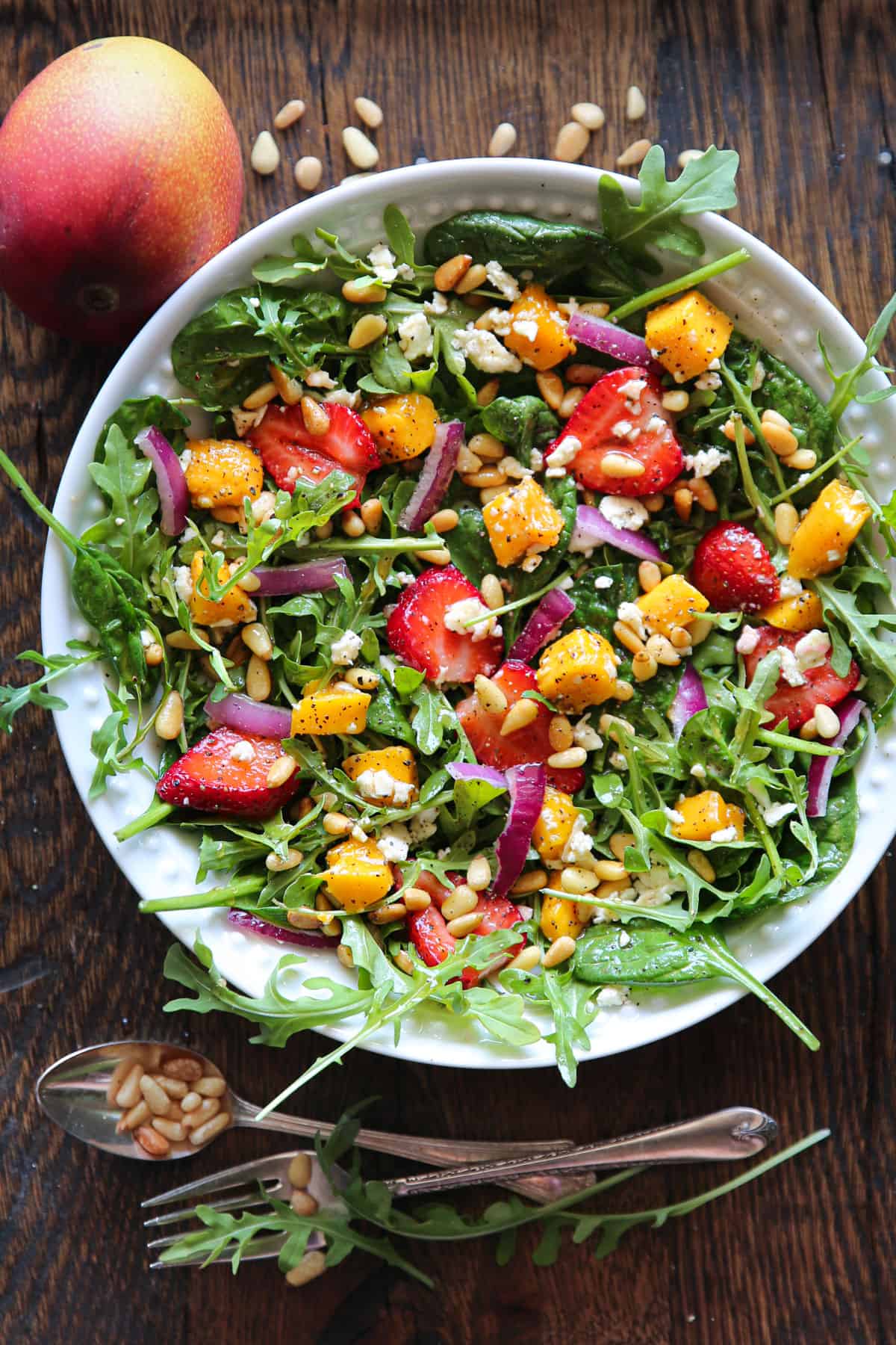 Strawberries and mango salad with fresh greens (arugula and spinach), sliced red onion, feta cheese, and pine nuts in a lemon honey mustard dressing - in a white bowl.