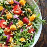 Strawberries and mango salad with fresh greens (arugula and spinach), sliced red onion, feta cheese, and pine nuts in a lemon honey mustard dressing - in a white bowl.
