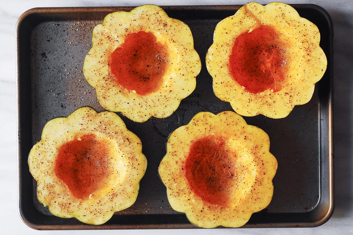 four acorn squash halves seasoned with olive oil, salt, and pepper on a baking sheet.