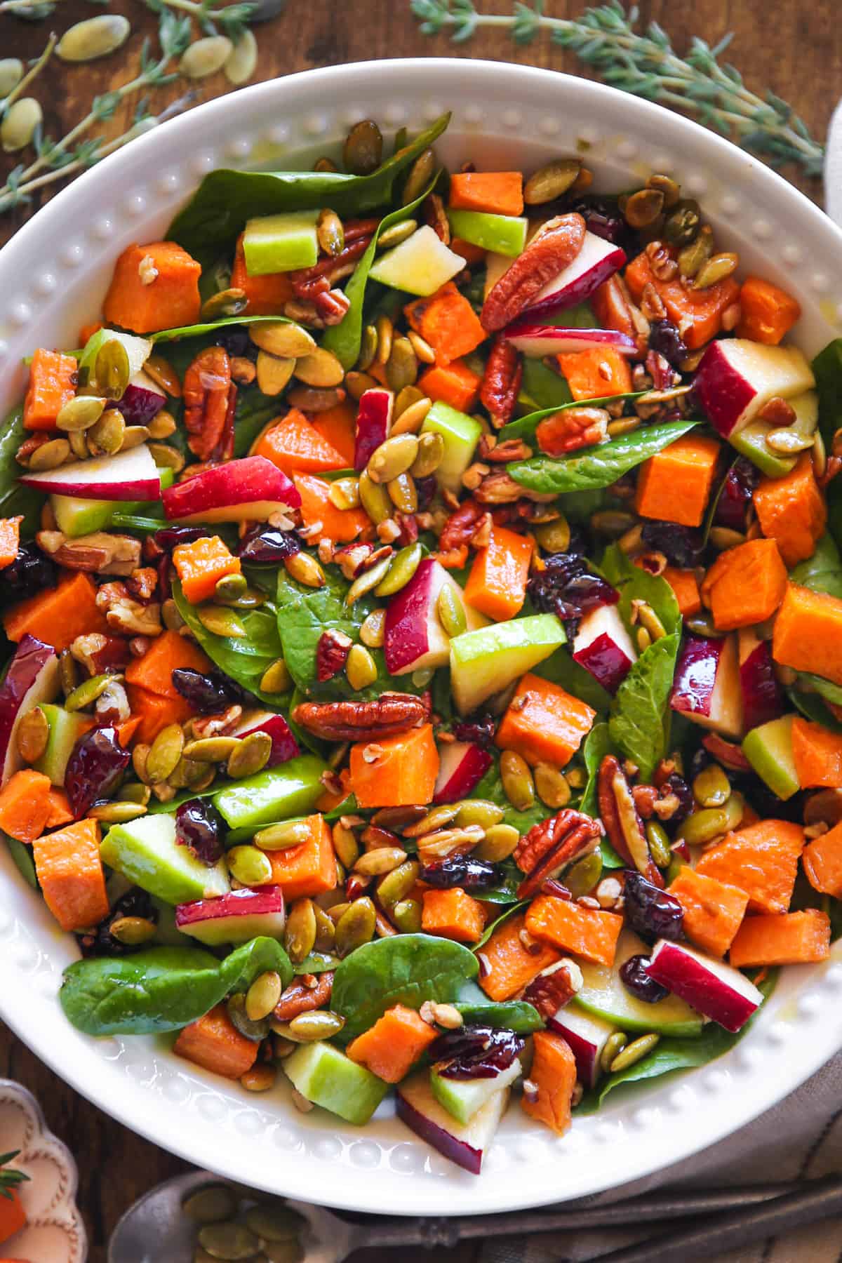 Roasted Sweet Potato Salad features spinach, apples, dried cranberries, pecans, pumpkin seeds in a white bowl.