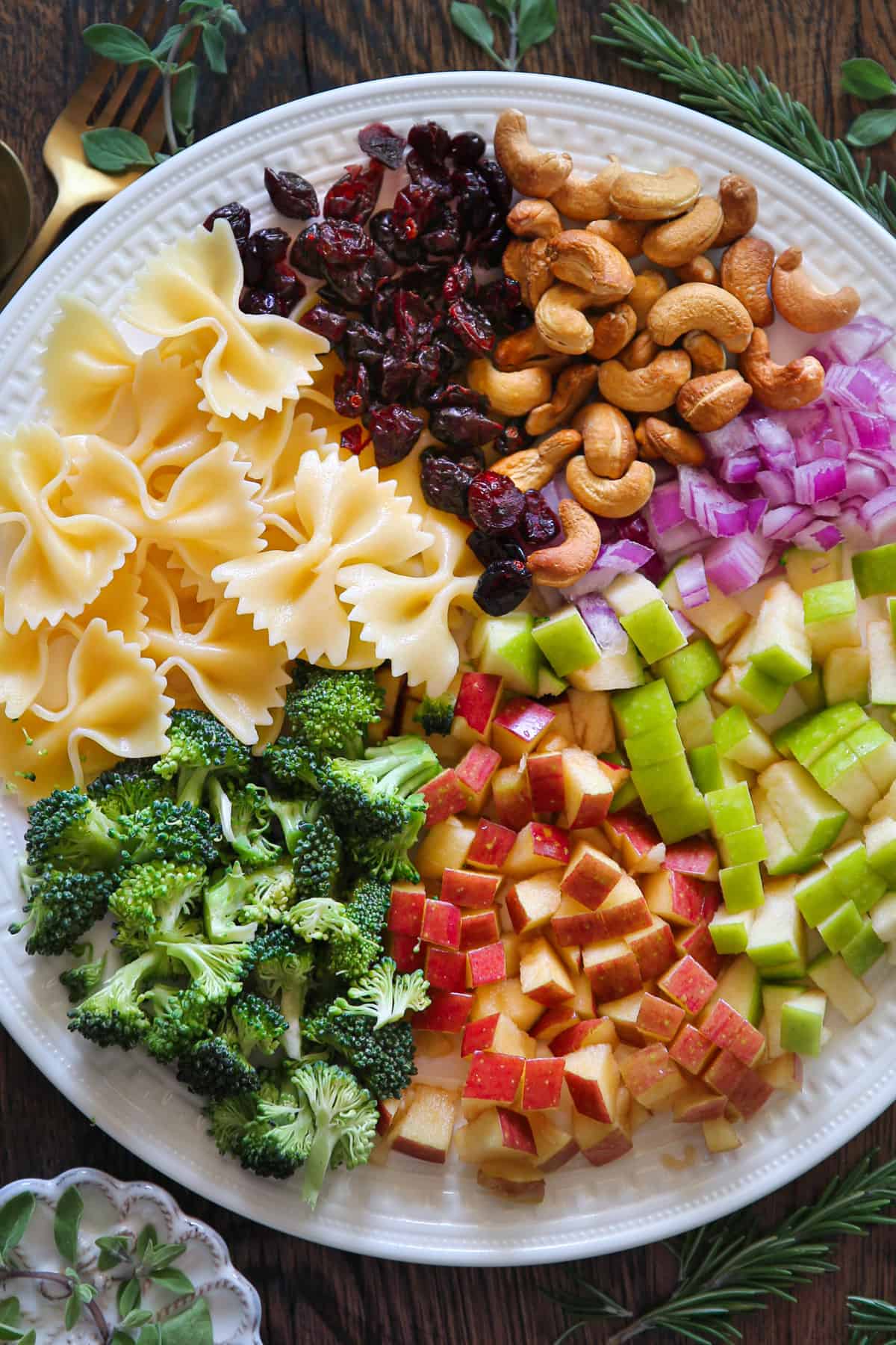 Zutaten für Brokkoli-Nudelsalat mit Preiselbeeren, Äpfeln, Cashewnüssen und roten Zwiebeln auf einem weißen Teller.