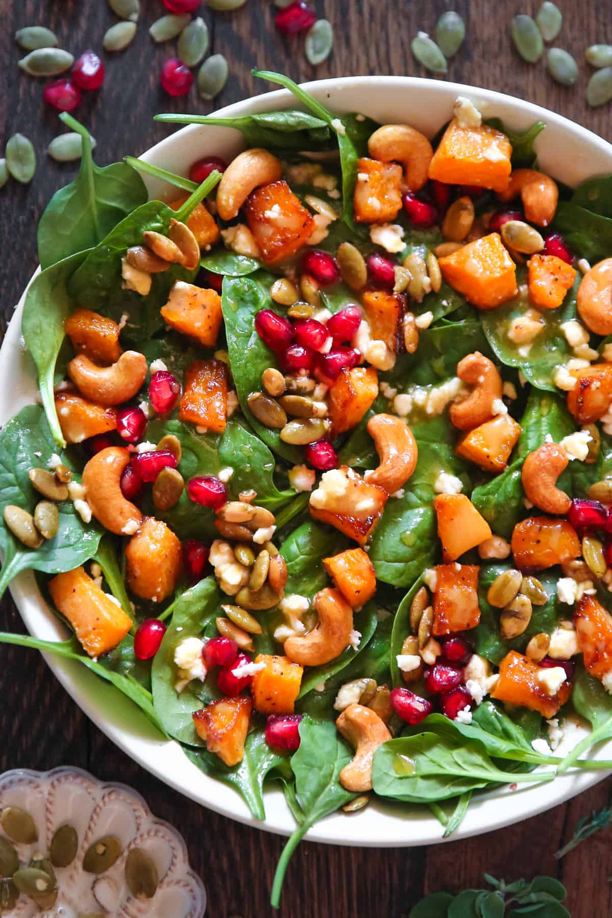 Butternut squash salad with spinach, cashews, pumpkin seeds, feta cheese, pomegranate seeds, and mustard honey-lime dressing in a white bowl.