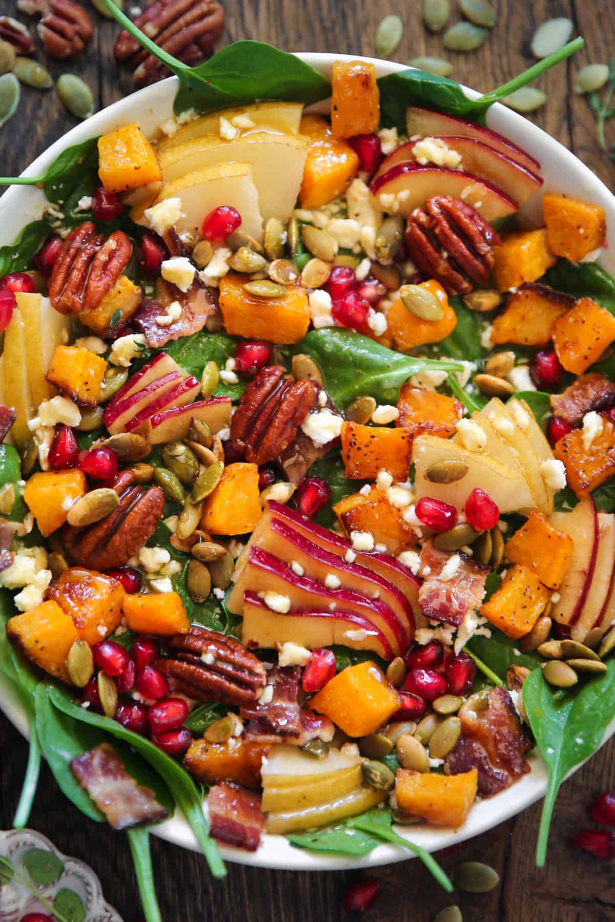 Fall Harvest Salad with Spinach, Butternut Squash, Apple, Pear, Pecans, Pumpkin Seeds, Bacon, Feta Cheese, Pomegranate Arils in a white bowl.