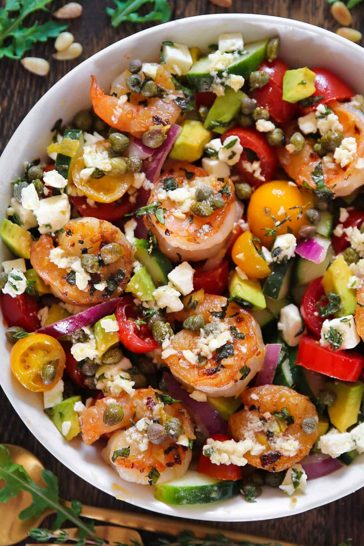 Greek shrimp salad wtih cherry tomatoes, cucumber, red onions, feta cheese, avocado, and capers - in a white bowl.