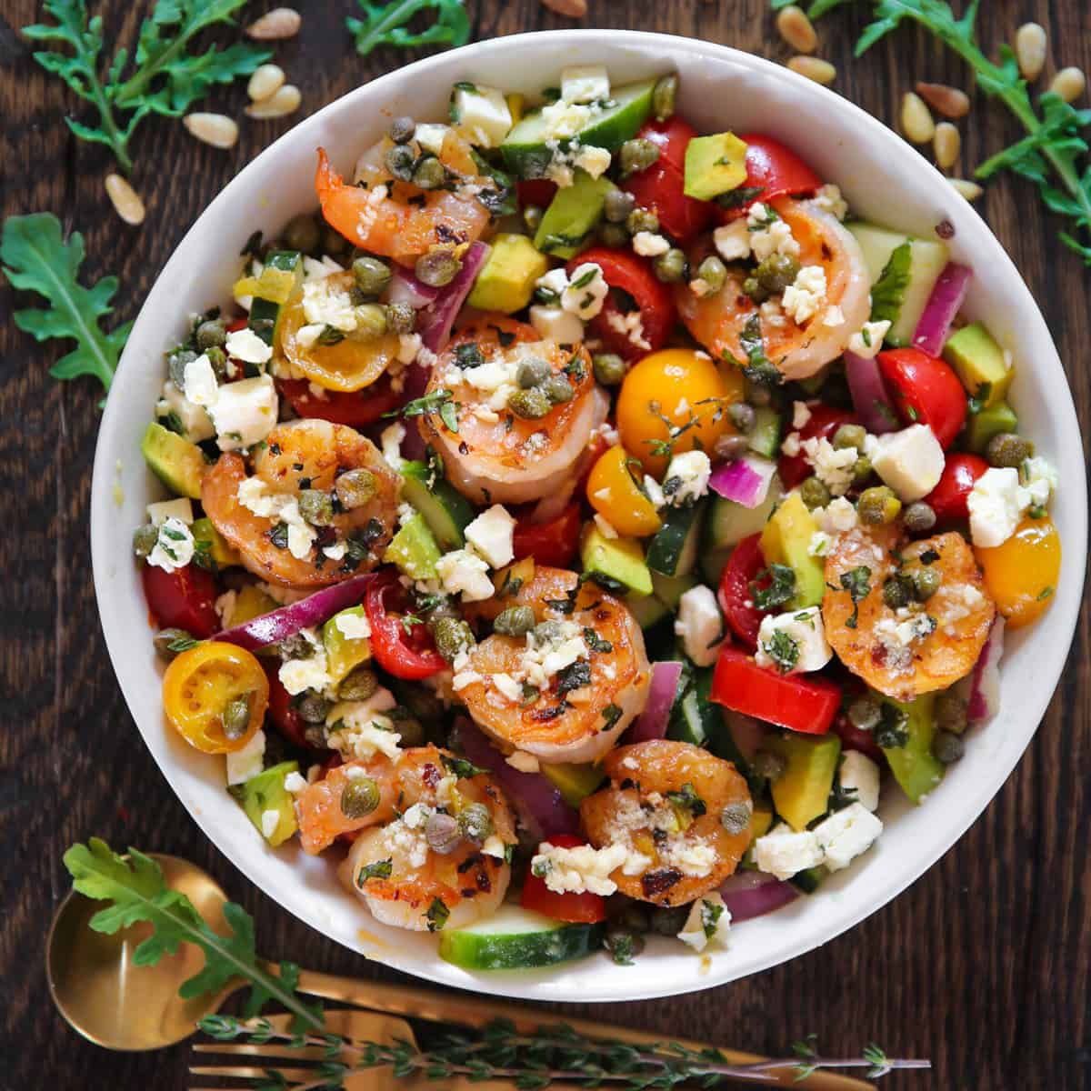 Greek shrimp salad wtih cherry tomatoes, cucumber, red onions, feta cheese, avocado, and capers - in a white bowl.
