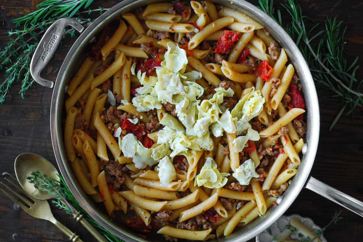 Italian sausage pasta with sun-dried tomatoes and artichoke hearts in a stainless steel skillet.