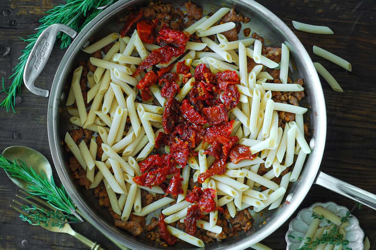 cooked sausage, uncooked rigatoni, and sun-dried tomatoes in a stainless steel skillet.