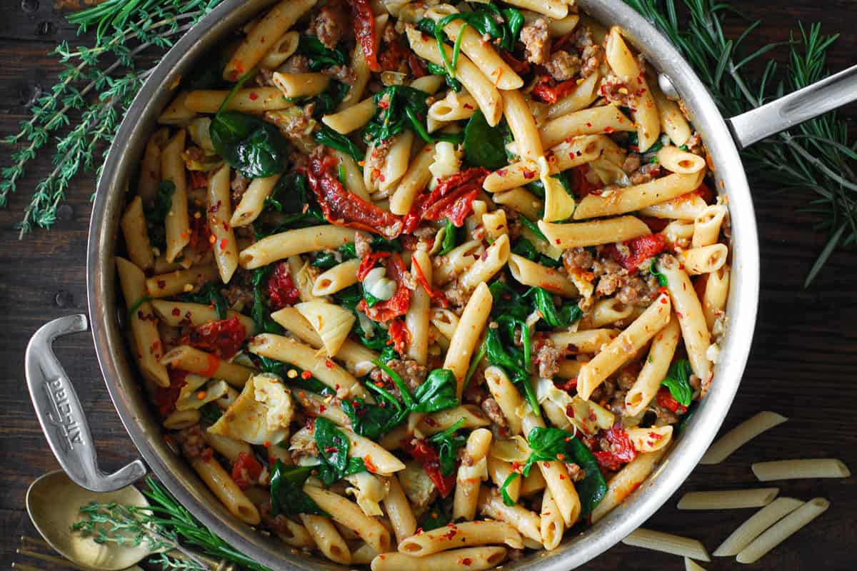 italian sausage rigatoni pasta with spinach, artichoke hearts, and sun-dried tomatoes - in a stainless steel skillet.