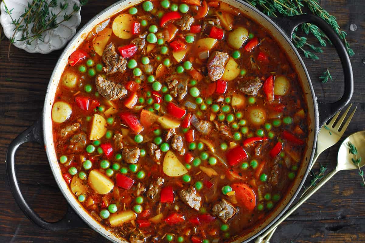 beef vegetable soup with onions, garlic, tomato paste, beef broth, sliced carrots, potatoes, bell peppers, and green peas - in a pot.