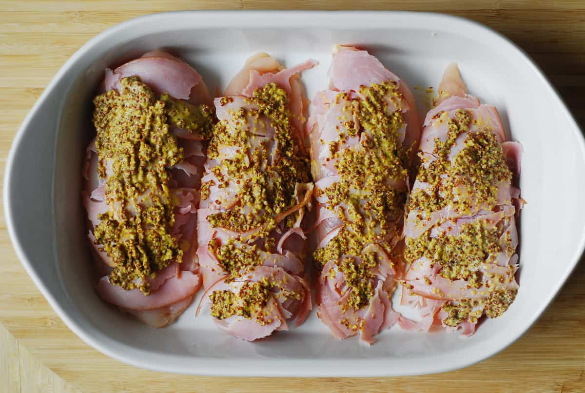 four chicken breasts with sliced ham and mustard in a casserole dish.