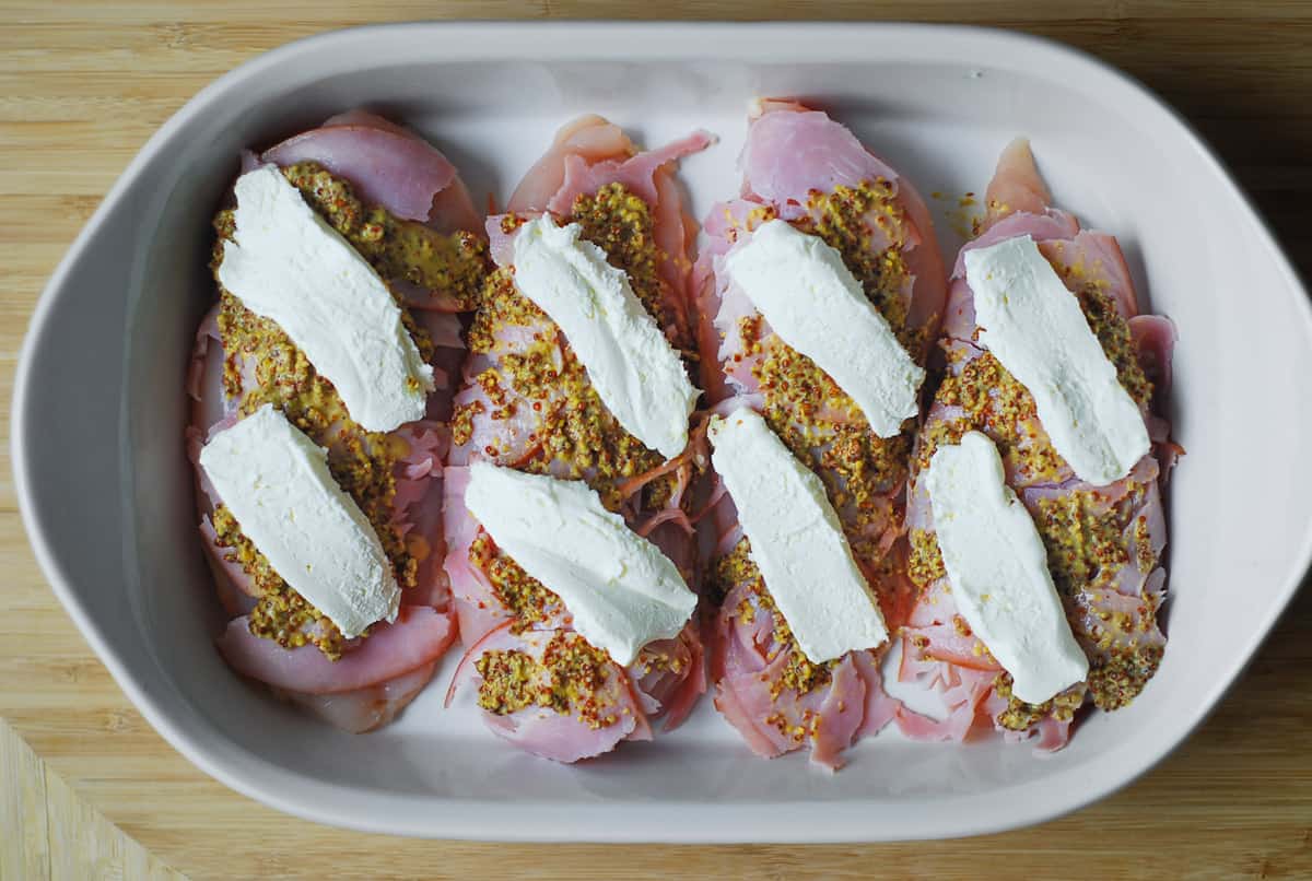 four chicken breasts with sliced ham, mustard, cream cheese in a casserole dish.