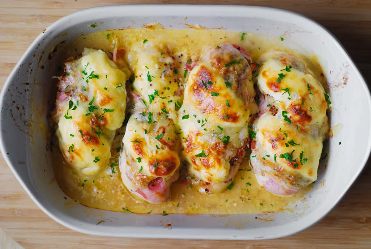 four cooked chicken breasts with sliced ham, mustard, cream cheese, Swiss cheese in a casserole dish.