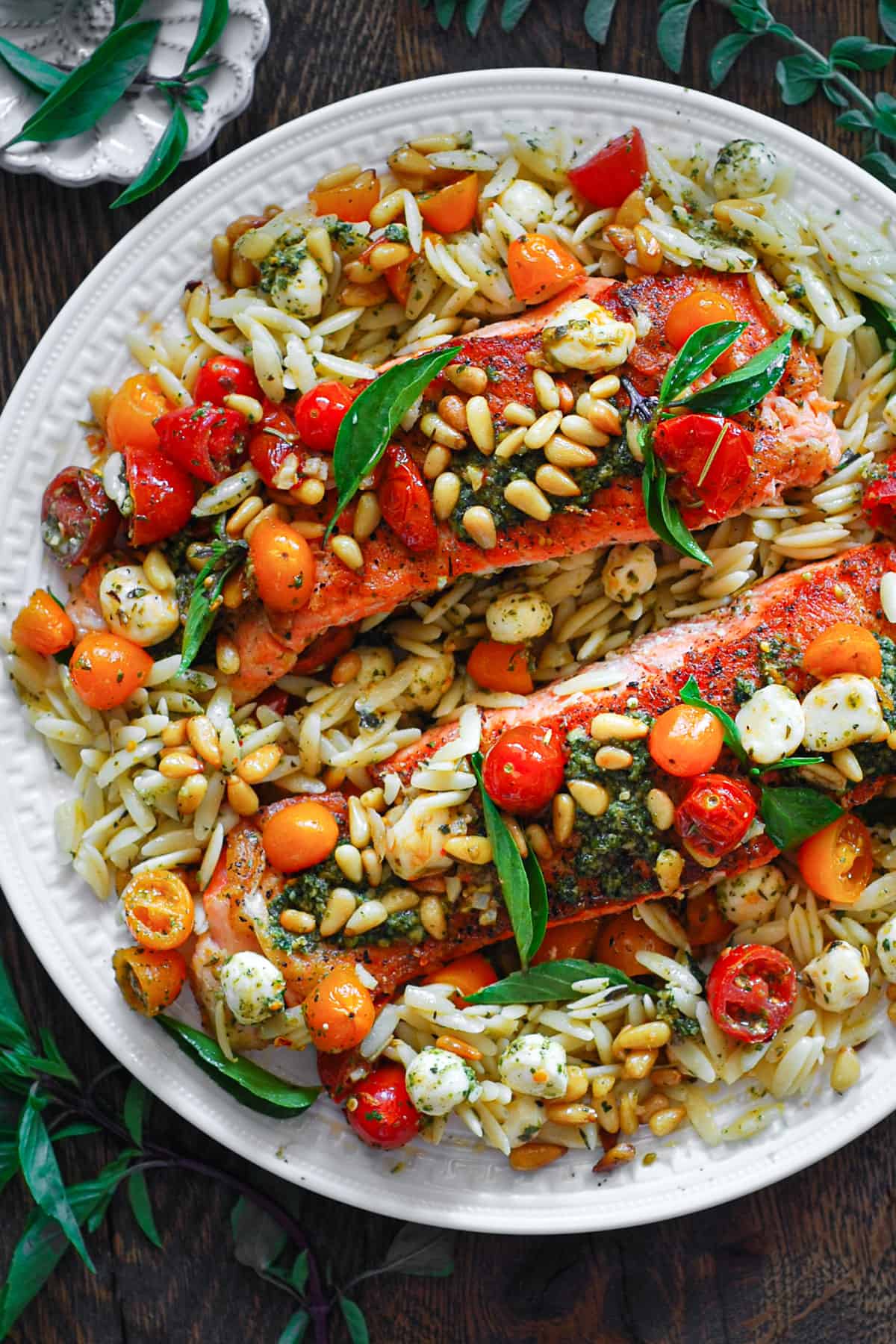 Salmon fillets with orzo, basil pesto, grape tomatoes, Mozzarella cheese, and pine nuts - on a large white plate.