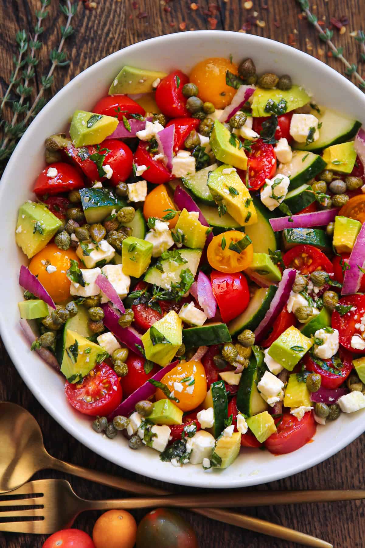 Greek Salad with tomatoes, cucumber, red onions, avocado, capers, and feta cheese - in a white bowl.