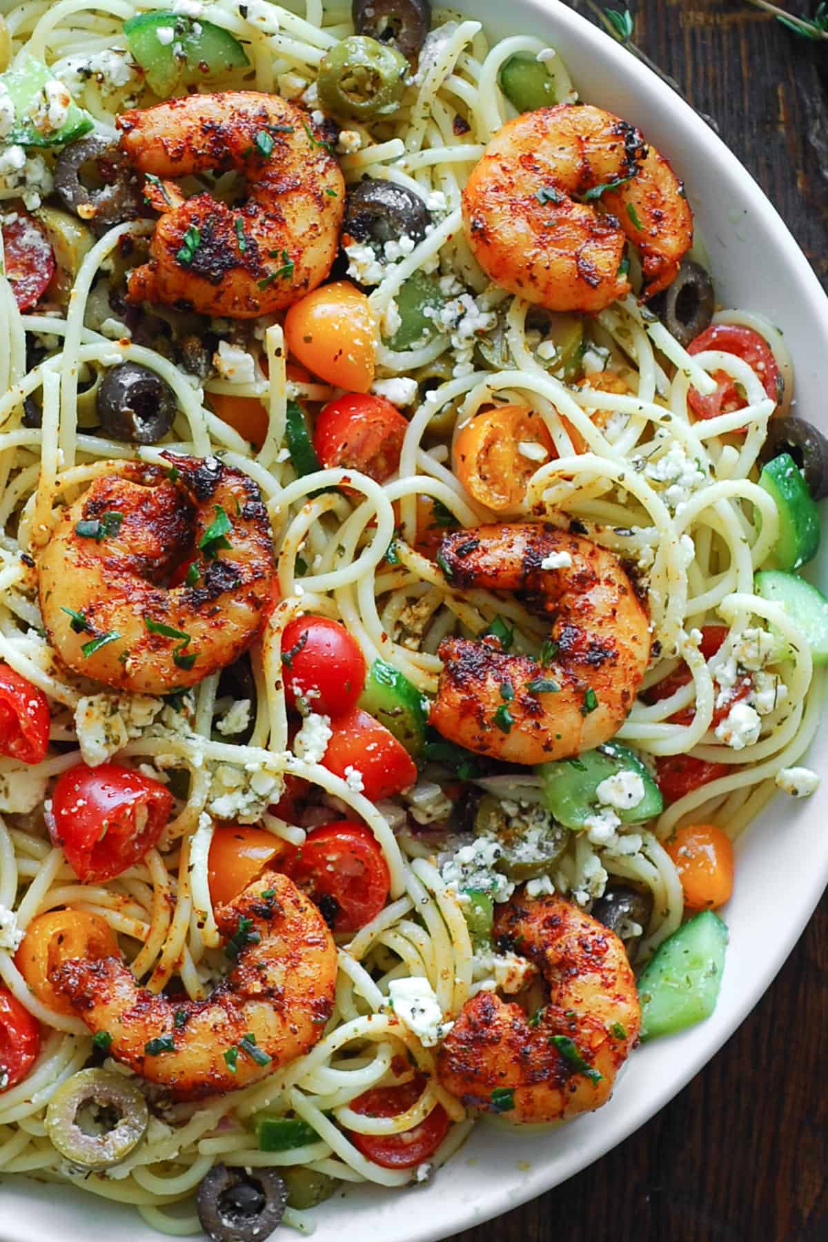 Garlic shrimp spaghetti with cherry tomatoes, olives, red onion, cucumber, feta cheese - in a white bowl. 