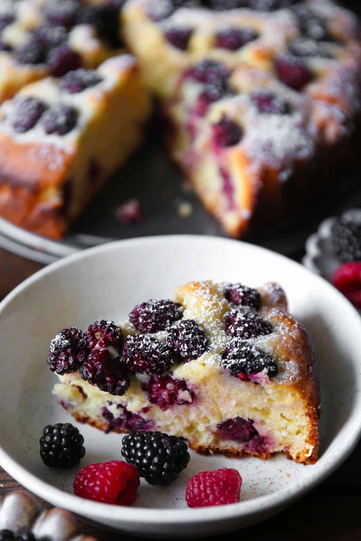 a slice of blackberry cake on a plate, with the remaining cake on a platter in a background.