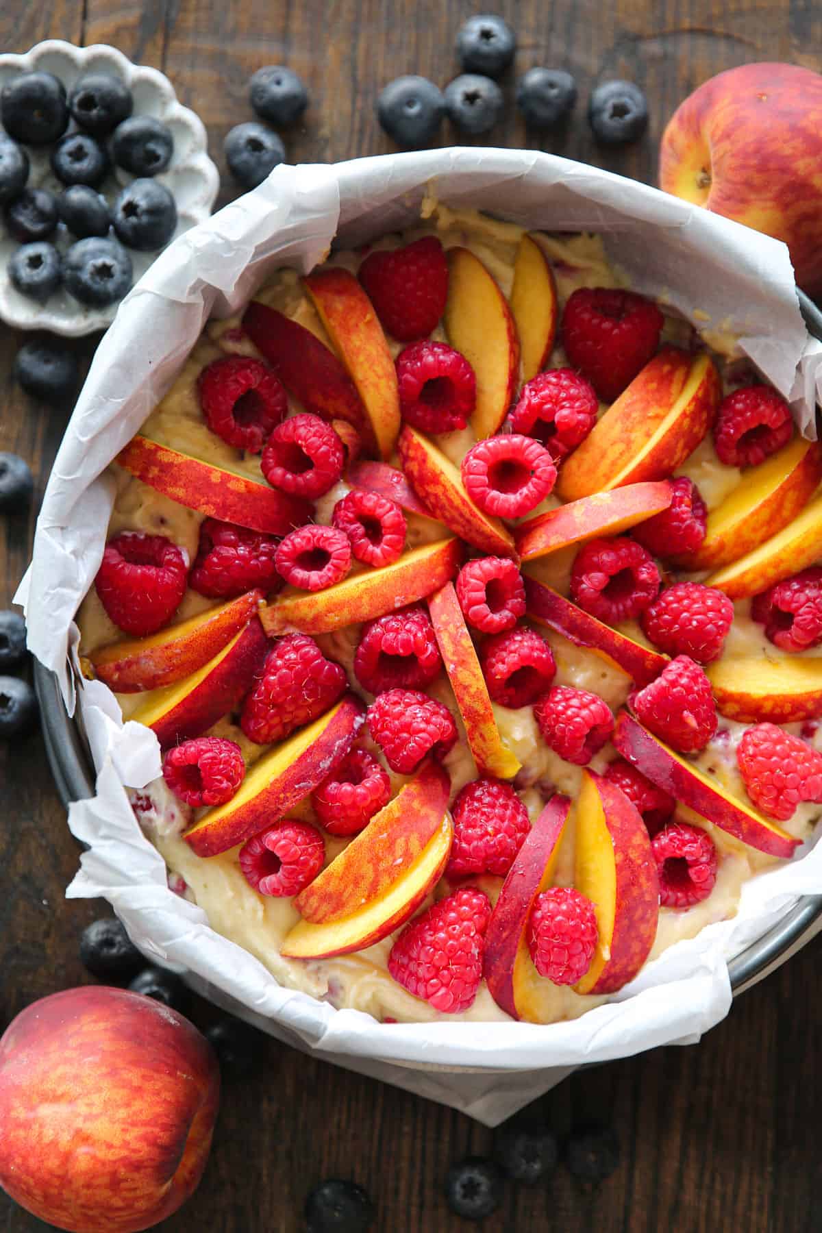 cake batter topped with sliced peaches and raspberries in a parchment paper-lined springform baking pan.