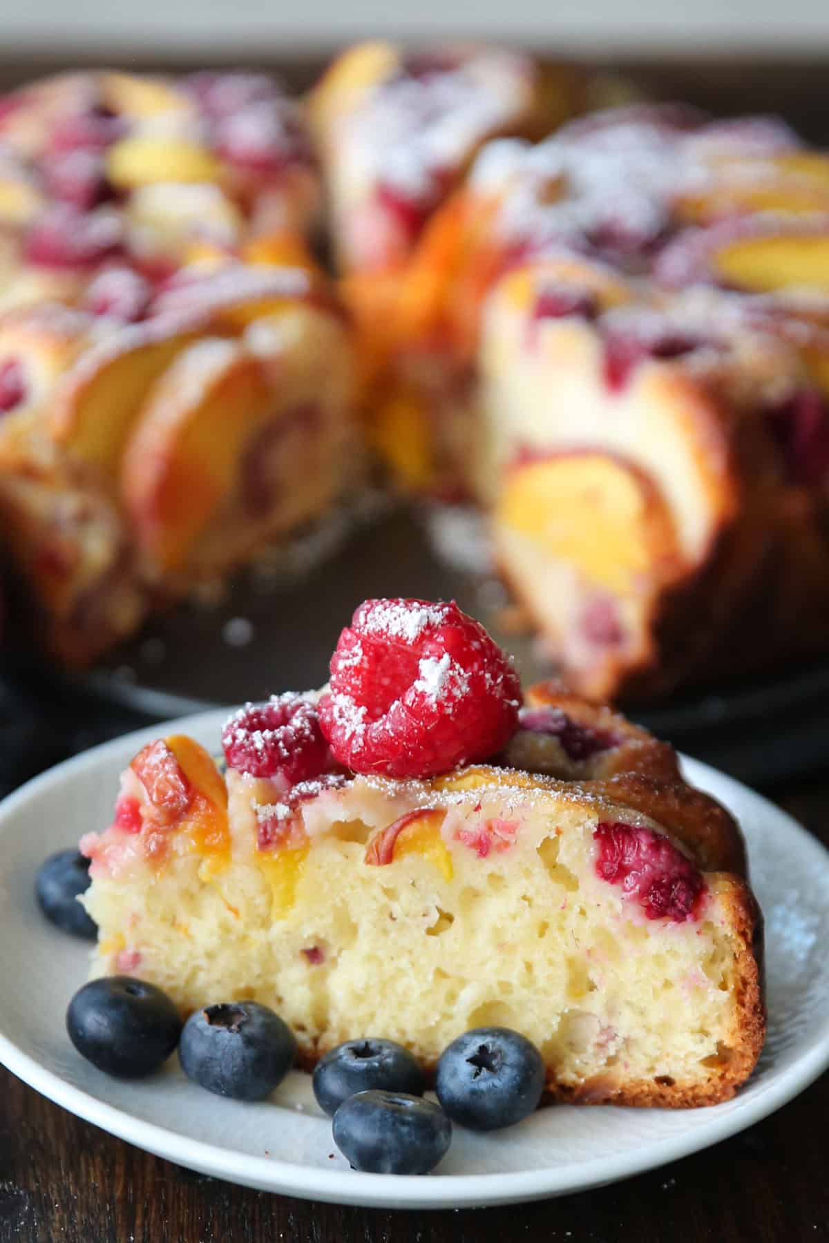 a slice of raspberry peach cake on a plate, with the whole cake in the background.