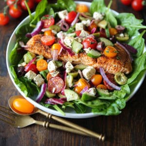 Greek salmon salad with spinach, kalamata olives, green olives, cherry tomatoes, cucumber, red onion, and feta - in a white bowl.
