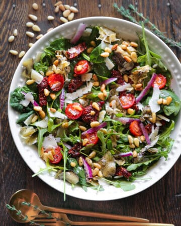 Italian Salad with spinach, arugula, sun-dried tomatoes, artichokes, cherry tomatoes, pine nuts, Parmesan cheese - in a white bowl.
