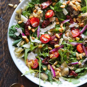 Italian Salad with spinach, arugula, sun-dried tomatoes, artichokes, cherry tomatoes, pine nuts, Parmesan cheese - in a white bowl.