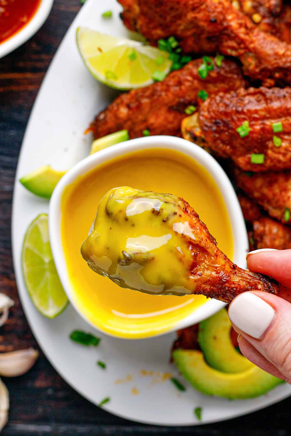 A hand holding a saucy chicken drumette dipped in yellow mustard sauce, with more chicken wings, lime wedges, and sliced avocado on a white plate in the background.